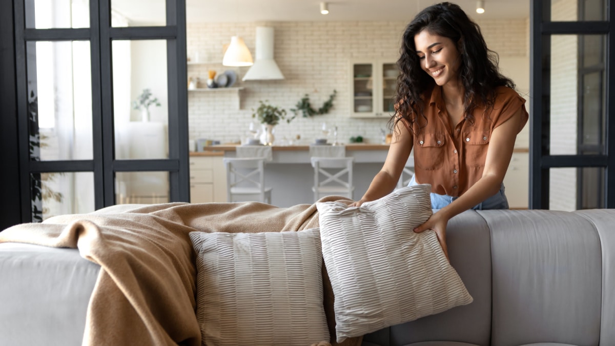 Cheap home decor - Lovely young woman putting soft pillows and plaid on comfy sofa, making her home cozy and warm, copy space.