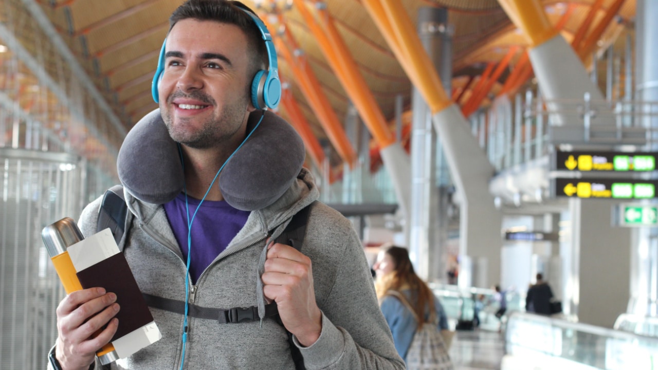 Happy man at airport with neck pillow, earphones, passport and tickets