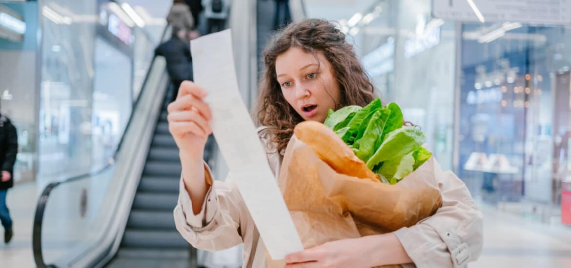 A woman in shock looks into a paper check from a supermarket in a shopping center against the background of an escalator and holds a package with fresh products, price increase
