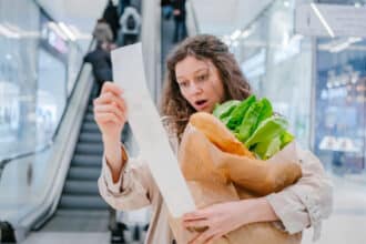 A woman in shock looks into a paper check from a supermarket in a shopping center against the background of an escalator and holds a package with fresh products, price increase