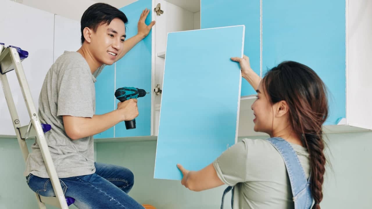 Cheerful young woman giving blue door to her husband assembling kitchen cupboard
