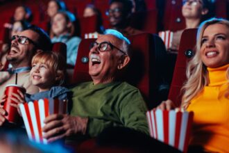 A crowd of happy spectators are in the movie, sitting in the chairs laughing at the movie they are watching and enjoying popcorn and drinks.