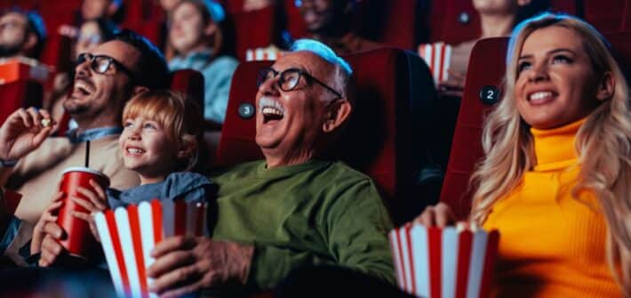 A crowd of happy spectators are in the movie, sitting in the chairs laughing at the movie they are watching and enjoying popcorn and drinks.