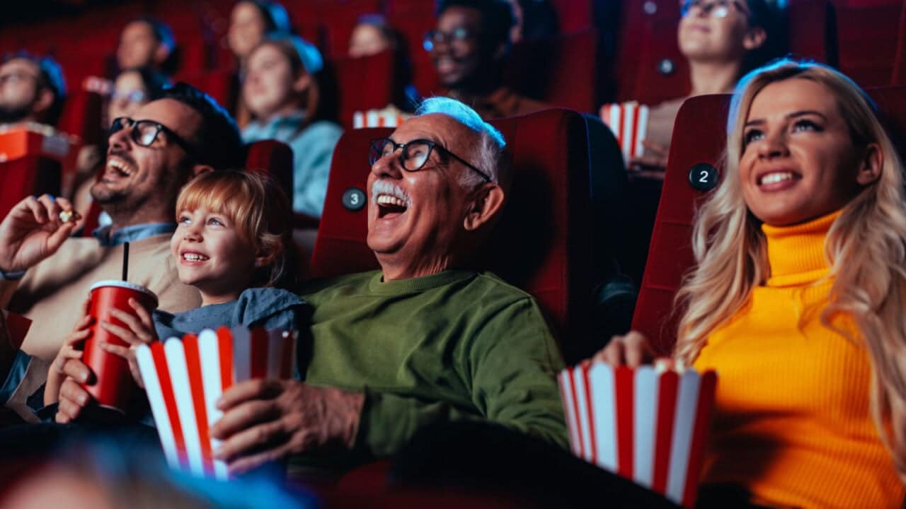 A crowd of happy spectators are in the movie, sitting in the chairs laughing at the movie they are watching and enjoying popcorn and drinks.