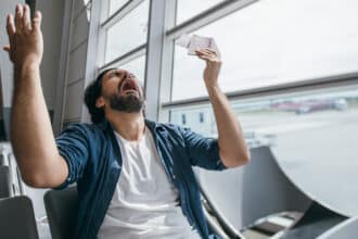 The man is very upset in the waiting room at the gate to the airport. A young guy, a traveler, waiting for a plane to fly out against the background of a large window, is very sad.