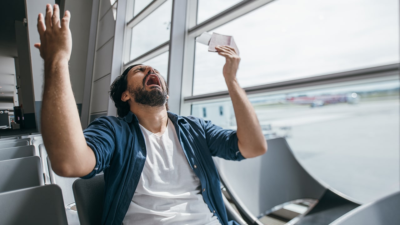 The man is very upset in the waiting room at the gate to the airport. A young guy, a traveler, waiting for a plane to fly out against the background of a large window, is very sad.