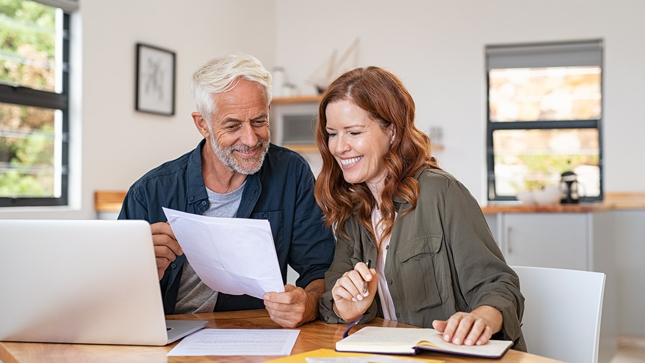 Mature smiling couple sitting and managing expenses at home. Happy senior man and mid woman paying bills and managing budget. Middle aged couple checking accountancy and bills while looking receipt.