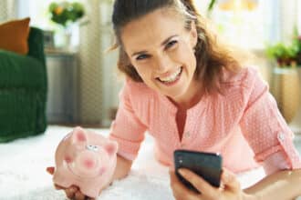 Portrait of smiling modern woman in blouse and white pants with piggy bank searching for carpet cleaning service on a smartphone while laying on white carpet at modern home in sunny day.