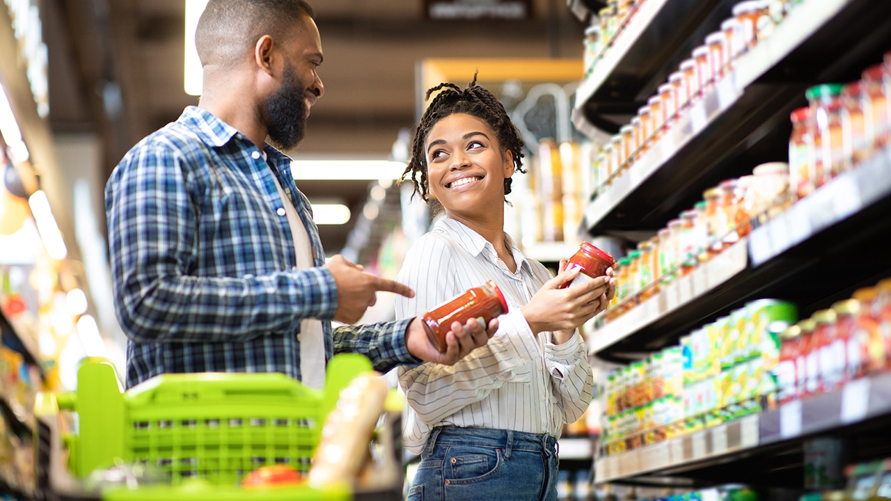 Happy African Family Couple Buying Food In Supermarket, Choosing Products Walking With Cart Along Aisles And Full Shelves Purchasing Groceries Together. Black Spouses Purchasing Essentials Together