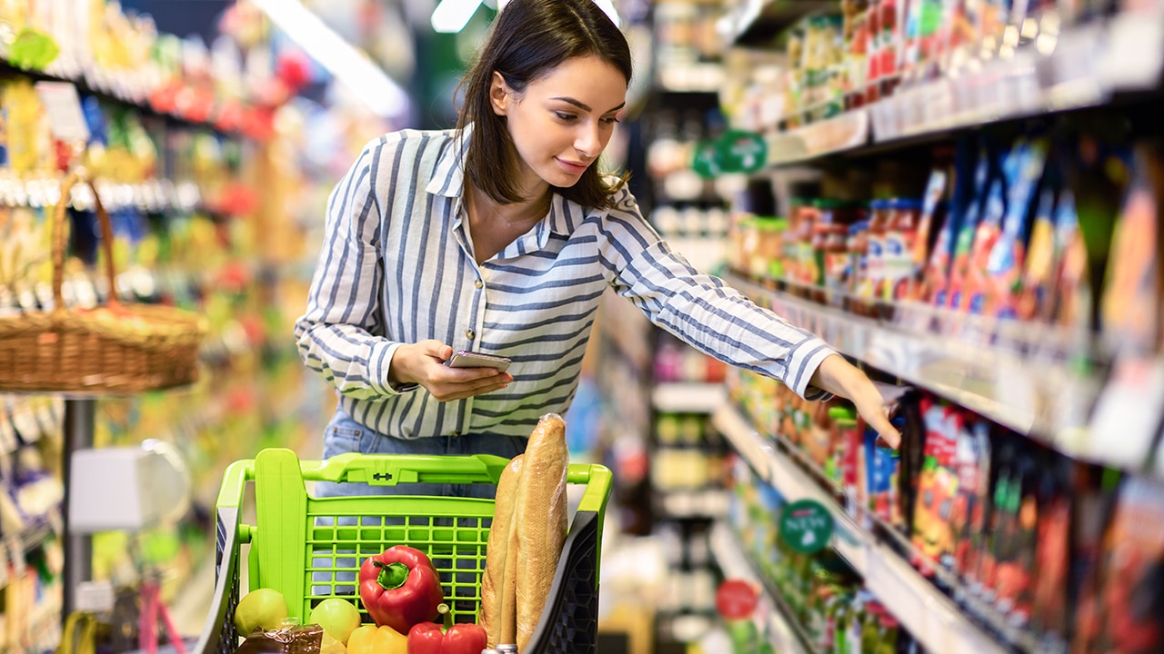 Portrait Of Millennial Lady Holding And Using Smartphone Buying Food Groceries Walking In Supermarket With Trolley Cart. Female Customer Shopping With Checklist, Taking Products From Shelf At The Shop