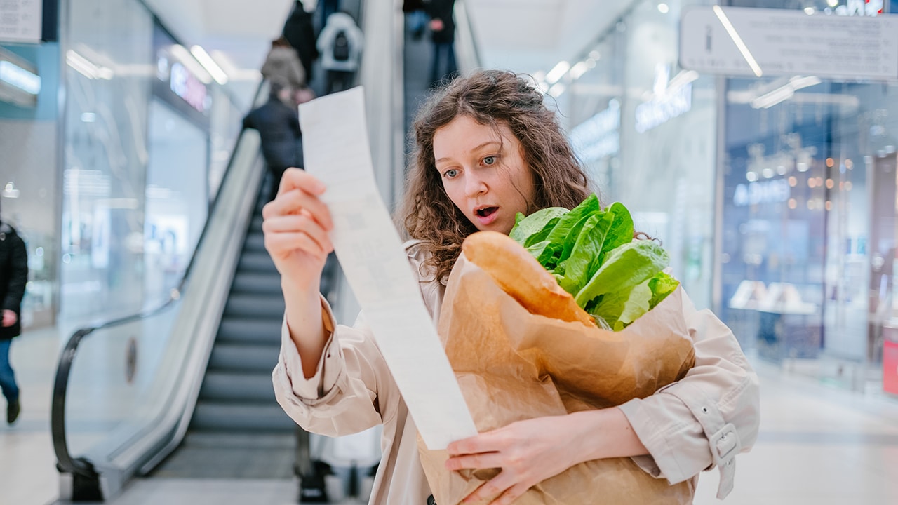 A woman in shock looks into a paper check from a supermarket in a shopping center against the background of an escalator and holds a package with fresh products, price increase