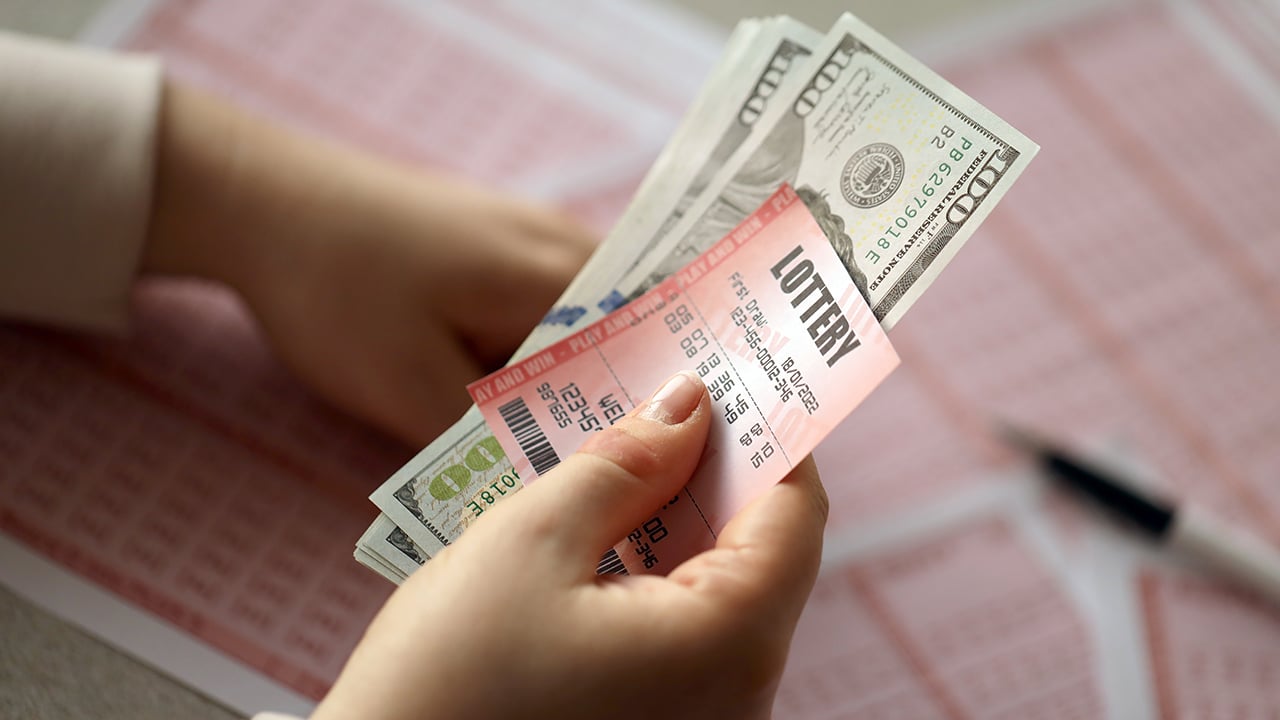 A young woman holds the lottery ticket with complete row of numbers and dollar bills on the lottery blank sheets background. Lottery winner concept