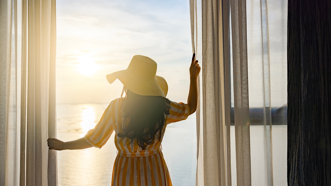 Rear back view woman opening curtains, relaxing and looking beautiful seascape panorama with rays of sun light in holiday on the balcony in high quality hotel feeling happy. Composition of nature