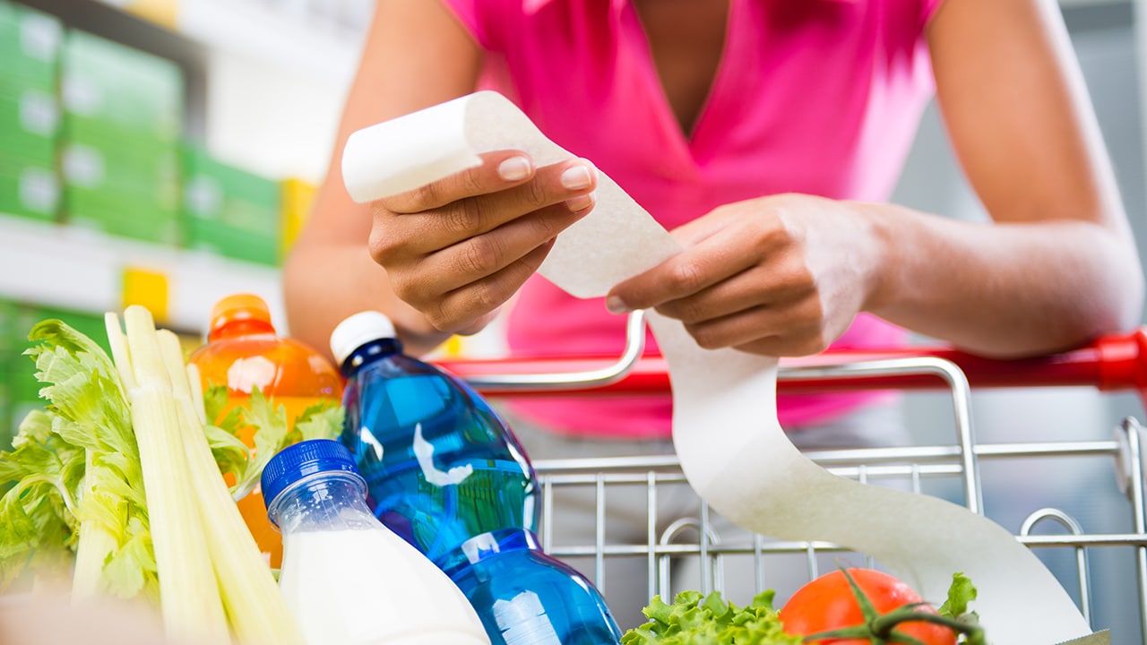 Unrecognizable woman checking a long grocery receipt leaning to a full shopping cart at store.