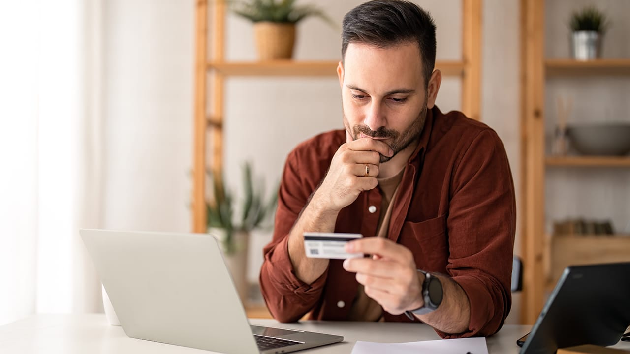 Serious business man sitting at table in home office, holding and looking at credit card, worried about debt and credit card deficit, considering a financial loan from bank.