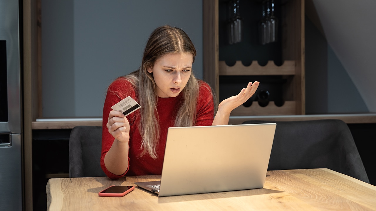 Young shocked frustrated woman holding bank credit card annoyed that she has reached her account limit and has no more money, she is refused online shopping and ordering on laptop computer