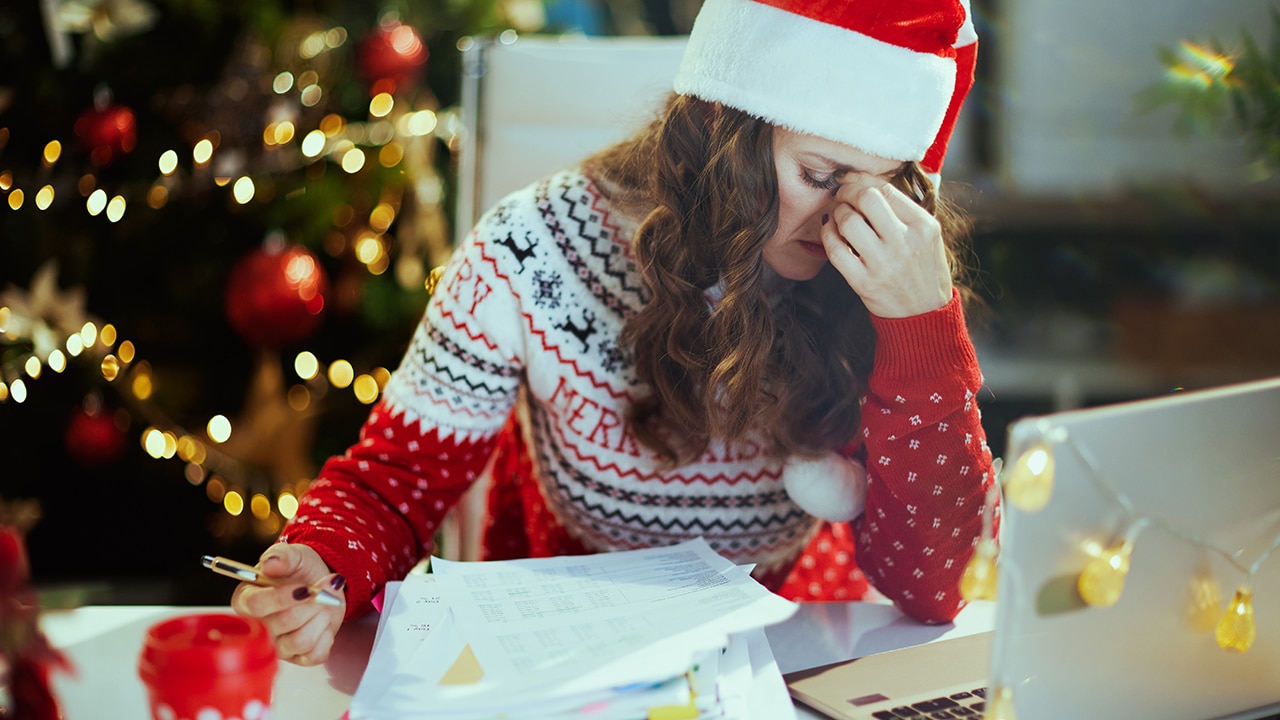 Christmas time. stressed stylish middle aged business woman in santa hat and red Christmas sweater with documents and laptop working in modern green office with Christmas tree.