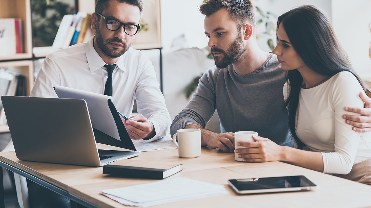 I need your signature here. Confident young man in shirt and tie holding some document and pointing it while sitting together with young couple at the desk in office