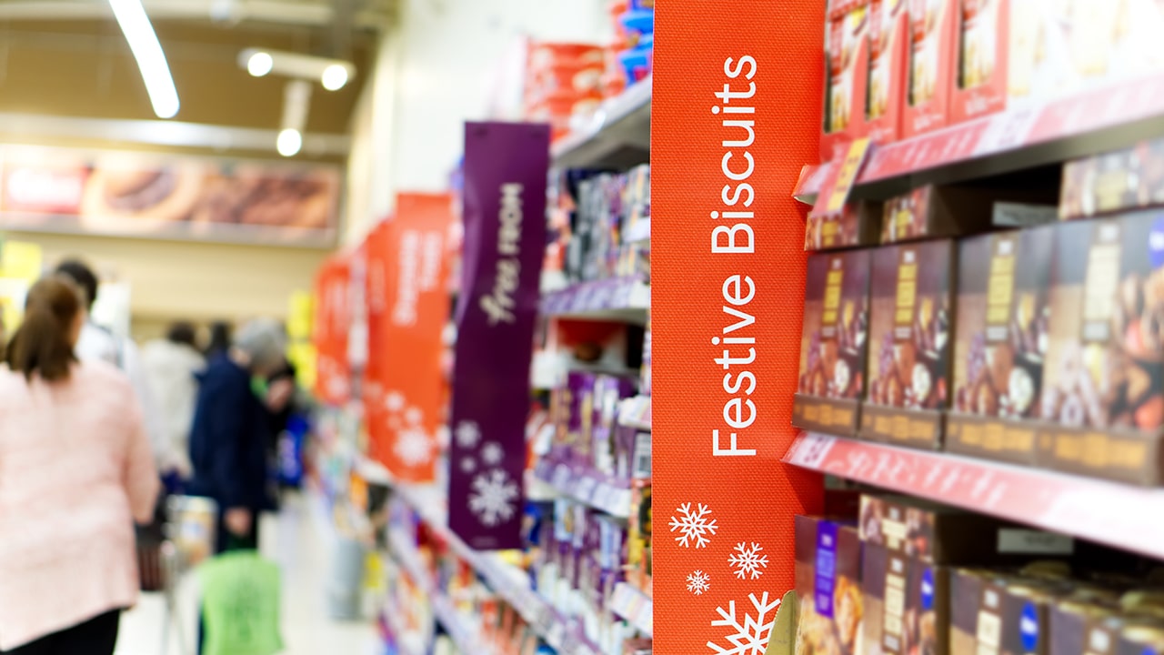 Christmas shoppers in a supermarket aisle with focus on a Festive Biscuits sign.