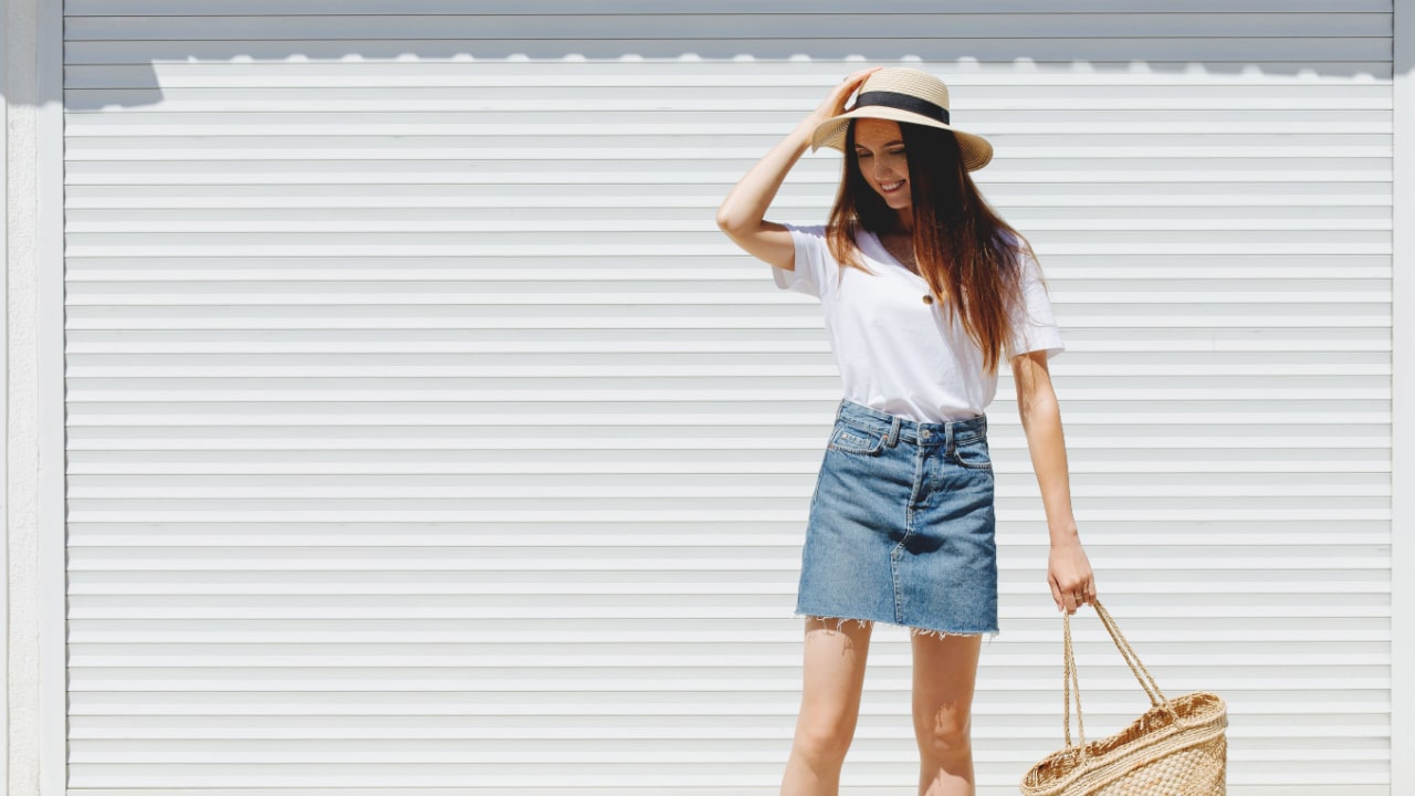 Young brunette woman wearing blue denim mini skirt, white t-shirt, wicker straw bag and boater hat.