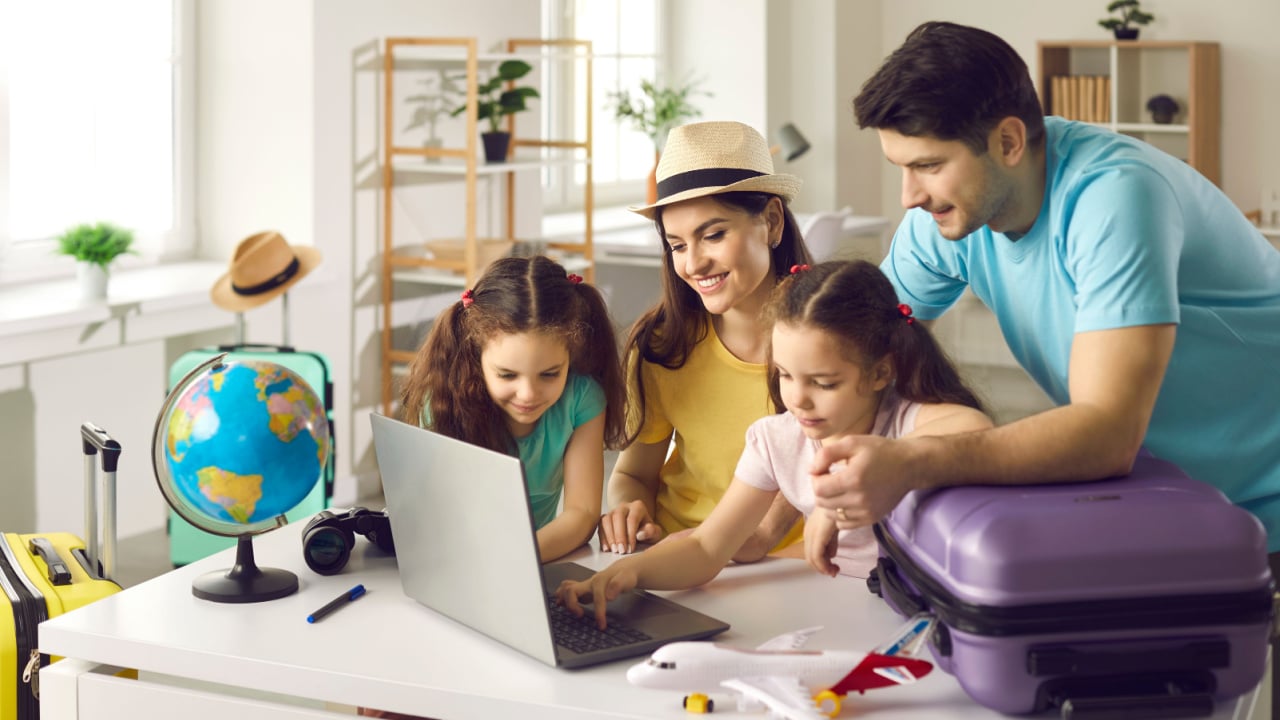 A family of man, woman and two girls looking at a laptop and a purple suitcase and toy plane on the desk