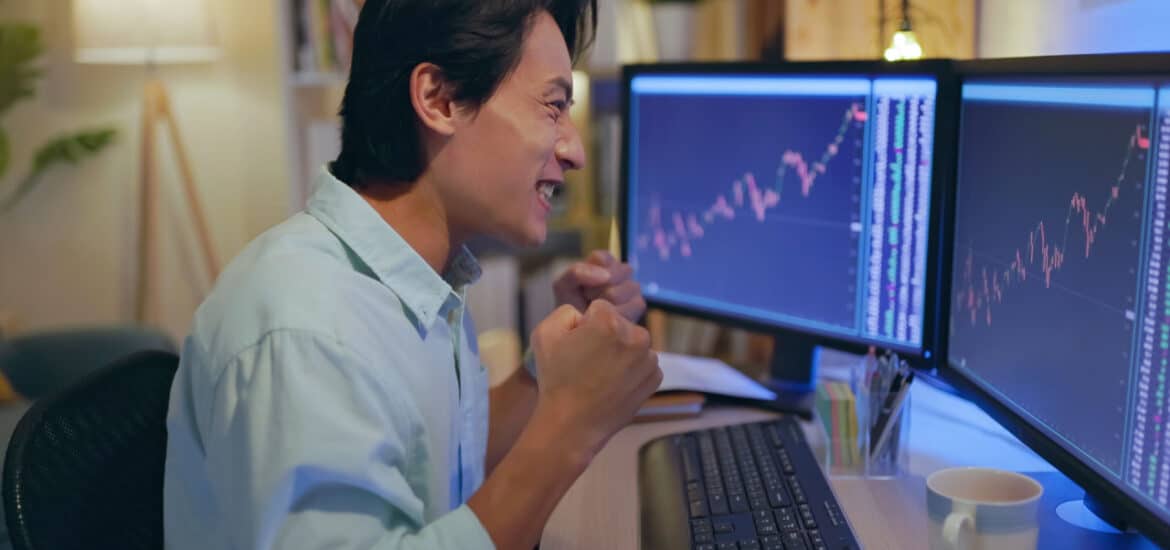 a happy man laughing and punching his fists looking at charts on two monitors