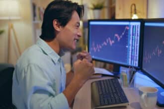 a happy man laughing and punching his fists looking at charts on two monitors