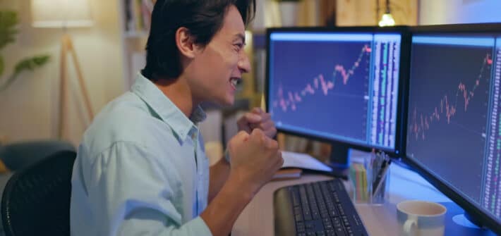 a happy man laughing and punching his fists looking at charts on two monitors