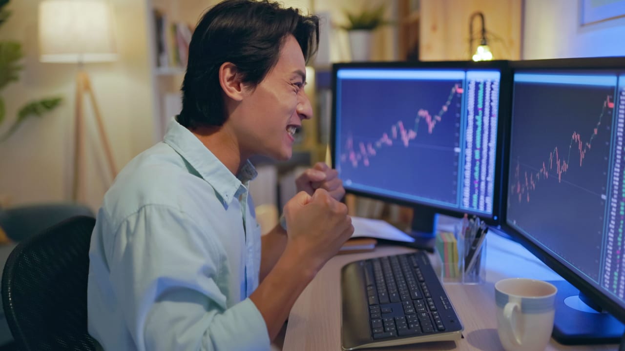 a happy man laughing and punching his fists looking at charts on two monitors