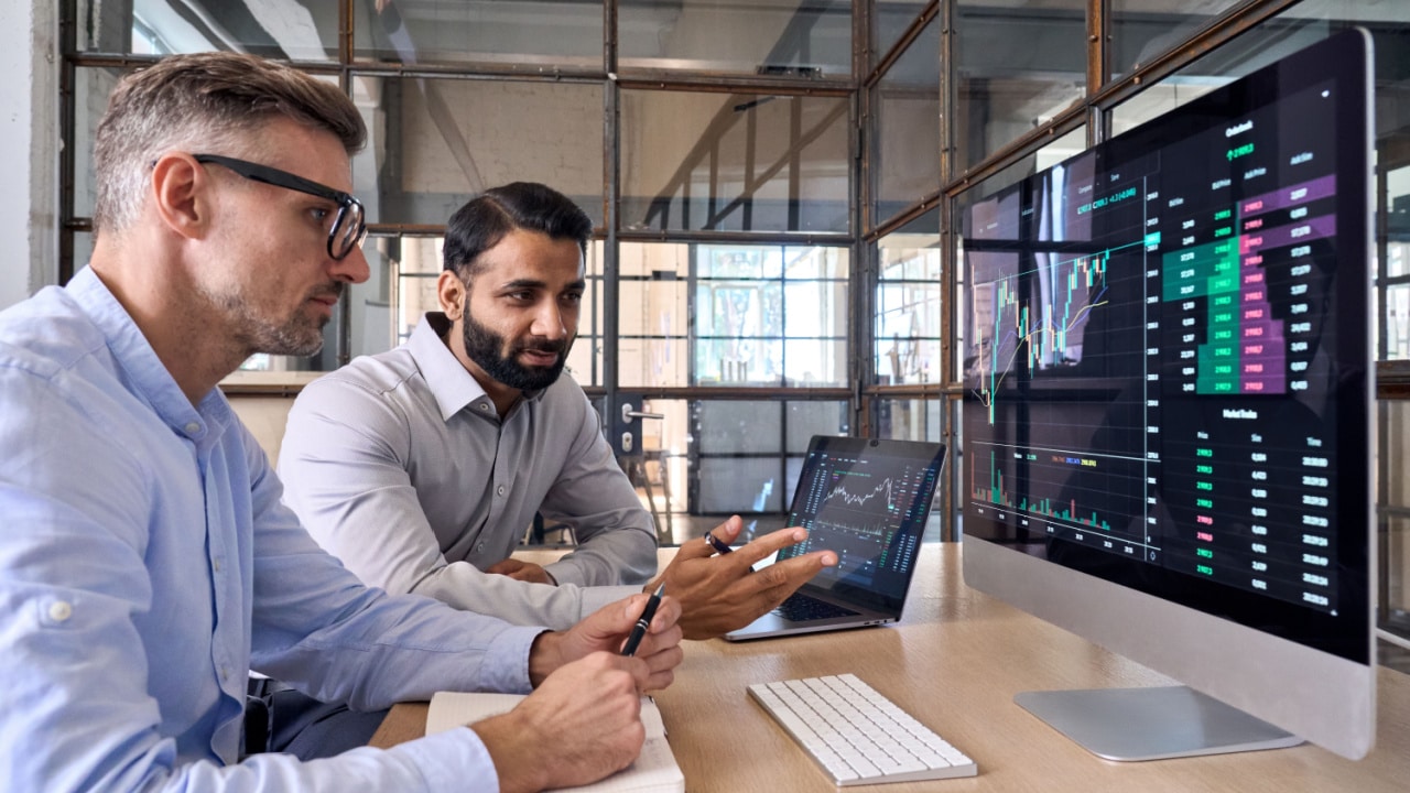 two men holding pens and looking at investment charts on a large monitor