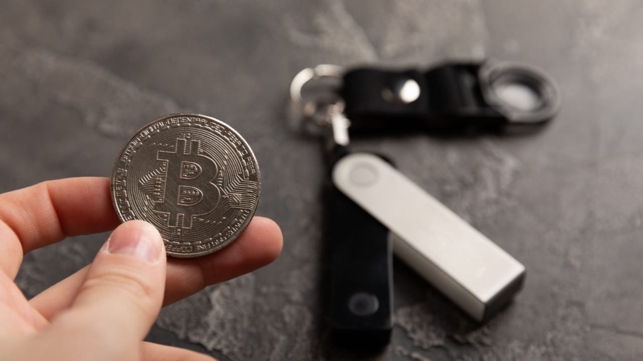 hand holding a bitcoin token with a cryptocurrency cold wallet in the background