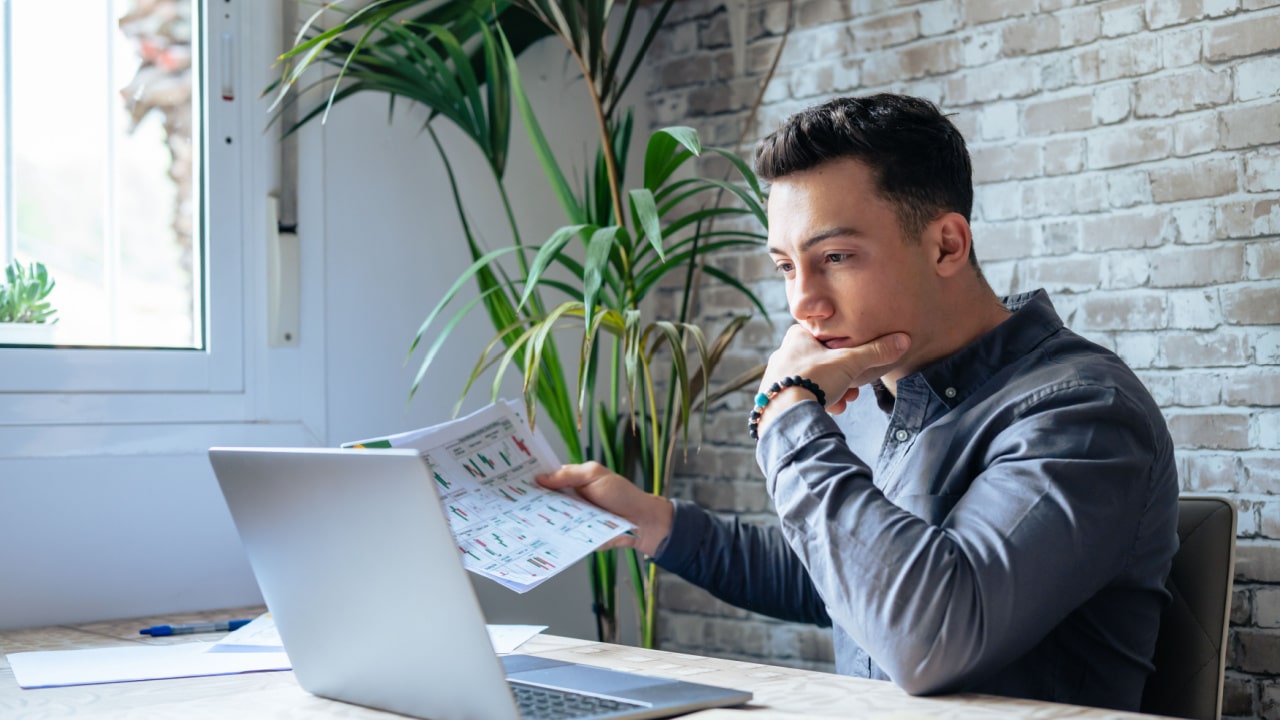 man looking at laptop and holding papers in his hand