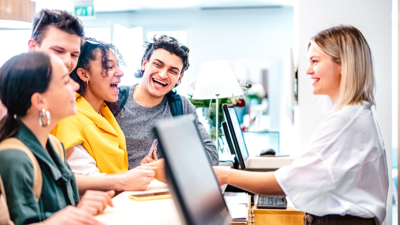 two young men and two young women laughing as they're being served by a female hotel receptionist