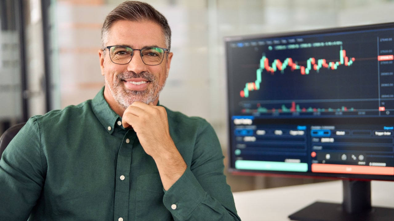 man smiling with a monitor in the background showing investment charts
