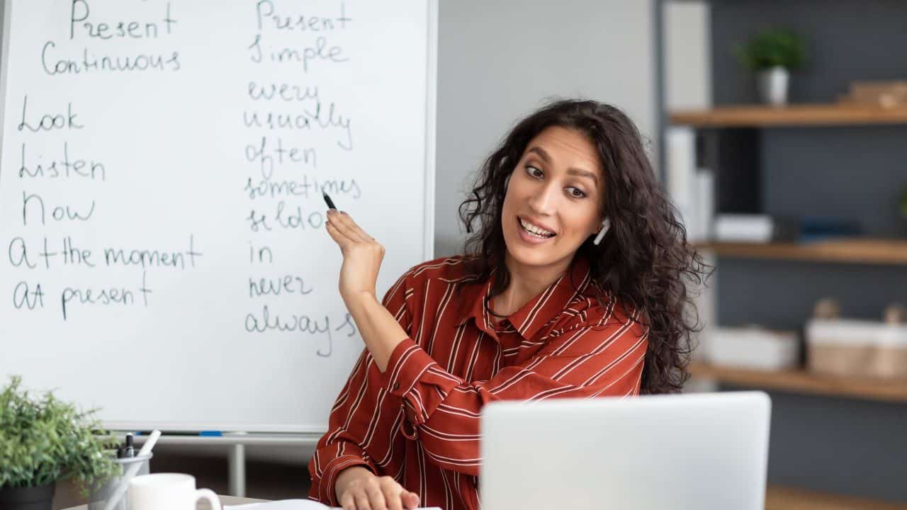 Online Foreign Languages Tutoring. Joyful female teacher giving English class, pointing at blackboard with basic grammar rules. Experienced college professor explaining new material tenses to students