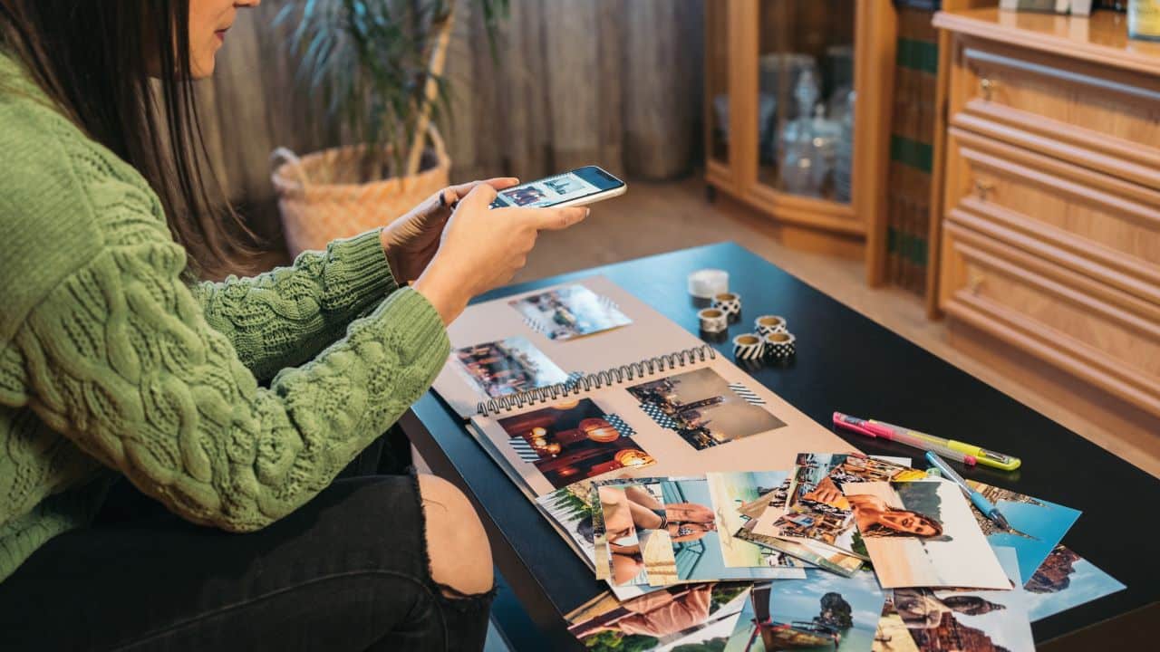 Middle-aged brunette woman sitting on her sofa taking a photo with her smartphone to the handmade kraft album with travel photos and washi tape.